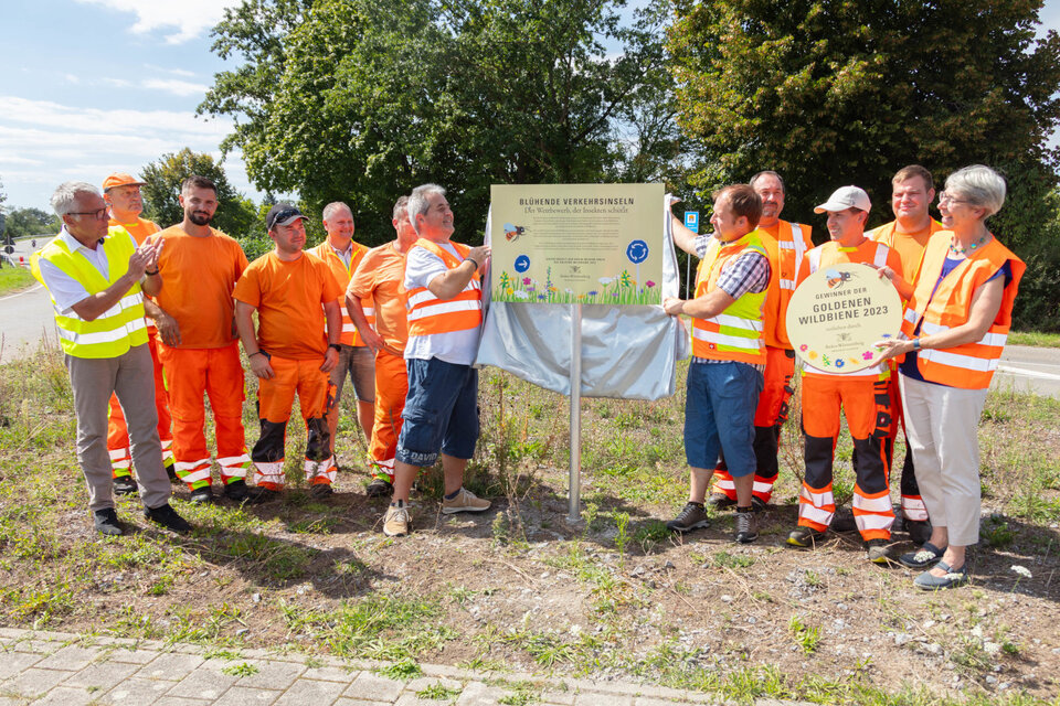 Rhein-Neckar-Kreis mit der „Goldenen Wildbiene“ ausgezeichnet: Landrat Stefan Dallinger (1.v.l), Staatssekretärin Elke Zimmer (1.v.r.), Verkehrsministerium Baden-Württemberg, enthüllen gemeinsam mit dem Team der Straßenmeisterei Weinheim das Schild „Blühende Verkehrsinseln“.