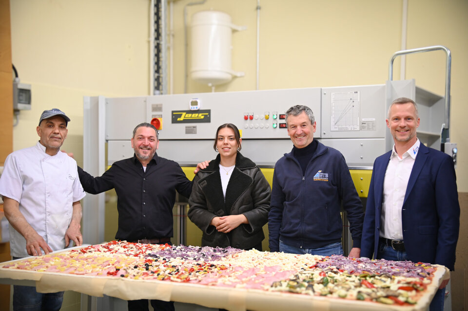 Handwerk trifft Kulinarik in der Gewerblichen Schule Tauberbischofsheim: Rocco Carella (2.v.l.) und sein Team von Carellas Ristorante versorgten die Teilnehmer mit einer einzigartigen Pizza. Ebenso einzigartig: Abteilungsleiter Jörg Schwab (v.r.), Schulleiter Hermann Ruppert und die Fachlehrer backten die 180 Zentimeter lange Pizza in der hauseigenen Furnierpresse der Schule. 